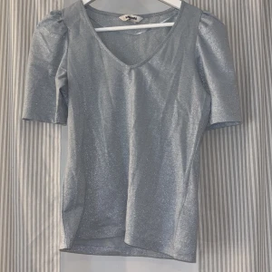 Glittrig silver/ljusblå topp från Kappahl - Glittrig topp från Kappahl i storlek S. Toppen puffärm och v-ringning. Materialet är stretchigt och glittrar i kontakt med lampor. Använd 1-2 gånger 