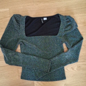 Glittrig långärmad topp från H&M Divided - Säljer en glittrig långärmad topp från H&M Divided i svart med inslag av grönt och blått. Toppen har fyrkantig urringning och lätt puffade axlar för en trendig look. Perfekt för dig som vill sticka ut lite extra.