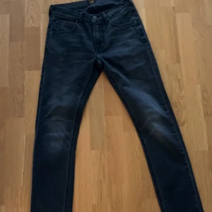 Mörkgrå slim jeans från Lee - Snygga mörkgrå jeans från Lee med klassisk femficksdesign och skinny passform. Jeansen har en stilren look med diskreta slitningar och Lee-logga på bakfickan. Perfekta för dig som gillar en smal siluett och tidlös stil.