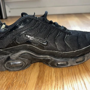 Nike Air Max Plus Tn helsvarta sneakers - Säljer ett par helsvarta Nike Air Max Plus Tn sneakers med mesh- och syntetöverdel, ikoniska vågformade detaljer och Tn Air-logga på hälen. Skorna har synlig Air-dämpning i sulan och klassisk snörning. Perfekta för dig som gillar streetwear och sportig stil.