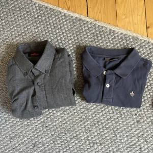 Två stilrena skjortor i grått och blått - Säljer två skjortor: en mörkgrå klassisk skjorta från Workers med knappar och krage, samt en marinblå piké med broderad detalj på bröstet. Båda har snygg passform och passar perfekt till jeans eller chinos.