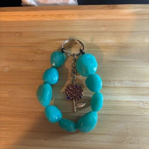 Turkost nyckelring med blomberlock - Stort armband med turkosa, oregelbundna pärlor och en söt blomberlock i lila och guld. Armbandet har en metallkedja och ring, perfekt för att ge din stil en färgklick och unik touch.