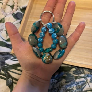 Turkost och grått nyckelring - Unikt armband med stora ovala pärlor i turkost och grått, blandat med mindre glittriga och mönstrade pärlor. Färgerna skiftar mellan blått, grått och inslag av beige. Perfekt accessoar för dig som gillar färg och statement-smycken.