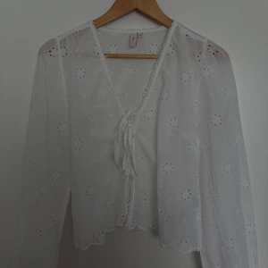 Vit blus med broderade blommor Nelly - Superfin vit blus från Nelly med broderade blommor och v-ringning. Blusen har långa ärmar, knytning framtill och fina virkade detaljer längs kanterna. 💛