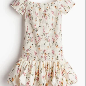 Blommig offshoulder kortklänning - Supersöt kortklänning med offshoulder och volangkant nertill. Klänningen är ljus med blommigt mönster i rosa, grönt och gult. Smockad överdel och korta ärmar ger en romantisk vibe, perfekt för varma dagar.