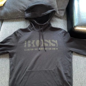 Svart hoodie från Hugo Boss - Svart hoodie från Hugo Boss i storlek S. Tröjan har en stor BOSS-logga framtill i ett diskret mönster, klassisk huva med snörning och en stor magficka. Perfekt för dig som gillar stilrena och bekväma plagg med premiumkänsla.