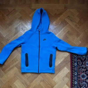 Blå Nike tech fleece med dragkedja - Nike hoodie i klarblå färg med svart logga på bröstet. Tröjan har hel dragkedja, två svarta fickor med dragkedja och en stor huva. Tillverkad i mjukt bomullsmaterial, perfekt för chill eller träning. Snygg och sportig look.