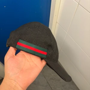 Svart Gucci keps med GG-mönster - Svart keps från Gucci med klassiskt GG-monogram över hela kepsen och grönt/rött band på sidorna. Justerbar rem baktill i svart med guldfärgat spänne. Snygg och stilren modell som ger en lyxig vibe.