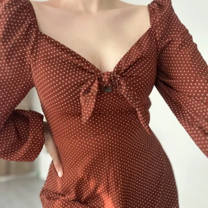 Brun prickig klänning med knyt - Superfin brun klänning med vita prickar, v-ringad och knyt framtill. Långa puffärmar och figurnära passform upptill, lätt utsvängd nertill. Perfekt för dig som gillar retrovibbar och söta detaljer. Passar S/M
