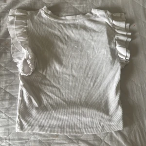 Vit ribbad topp med volangärmar - Supersöt vit ribbad topp med volangdetaljer på ärmarna. Den har en rund halsringning och är tillverkad i ett mjukt, stretchigt material som sitter bekvämt. Perfekt för dig som gillar en feminin och trendig stil.