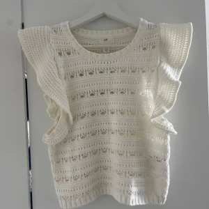 Vit stickad ärmlös topp H&M - Superfin vit stickad topp från H&M med breda volangdetaljer på axlarna. Den är ärmlös och har ett luftigt, virkat mönster som ger en riktigt somrig vibe. Perfekt att styla med jeans eller kjol för en fräsch look. Oanvänd💕💕