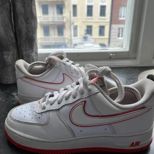 Nike Air Force 1 vita med röd sula - Nike Air Force 1 sneakers i vitt skinn med röda detaljer och röd yttersula. Klassisk låg modell med perforerad tå, snörning och tydlig röd Nike Swoosh-kant. AF1-märke på snörningen och röd logga på plösen. Perfekt för dig som gillar en clean men ändå unik look. Box medföljer även, några slitningar I skorna 