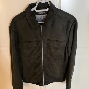 Human scales Jacka, Rick Vegan Suede - Snygg mörkgrön mockajacka från Human Scales med klassisk krage och två stora bröstfickor. Jackan har hel dragkedja framtill och är helfodrad med blått innerfoder. Perfekt för dig som vill ha en stilren och trendig look.