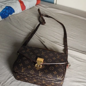  brun axelväska monogram - Säljer en klassisk brun axelväska  med ikoniskt monogrammönster och guldfärgade metalldetaljer. Väskan har justerbar axelrem, snygg låsdetalj framtill och dragkedja med gravyr. Perfekt för dig som gillar lyxig och tidlös stil.