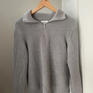 Grå stickad half zip-tröja East West - Säljer en grå stickad tröja från East West med half zip-dragkedja och hög krage. Tröjan är ribbstickad, har lång ärm och en clean, stilren look. Perfekt för lager-på-lager och chill vardagsstil.