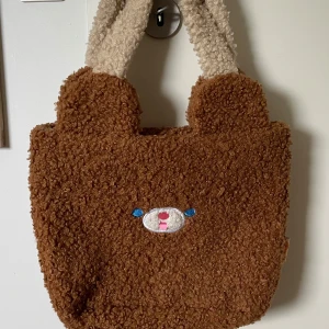 Brun teddy handväska från Milkjoy - Supergullig brun teddy handväska från Milkjoy med björnansikte broderat på framsidan och öron upptill. Väskan har fluffiga, ljusbeiga handtag och är perfekt för dig som gillar söta accessoarer med unik stil. Knappt använd!