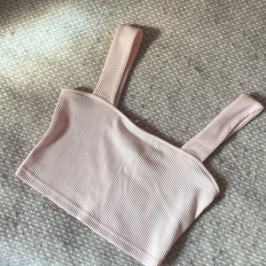 Ljusrosa ribbad linnetopp - Supersöt ljusrosa linnetopp med breda axelband och ribbad struktur. Toppen är croppad och har en enkel, stilren design som passar perfekt till jeans eller kjol. Materialet är mjukt och stretchigt för en bekväm passform.