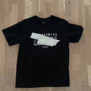 Maison Margiela svart t-shirt - Svart t-shirt från Maison Margiela med ikoniskt tryck av siffror och tejpdetalj framtill. Klassisk passform med rund hals och korta ärmar. Tillverkad i mjuk bomull och har märkets signaturdetaljer på ryggen. Perfekt för dig som gillar streetstyle och designerplagg.