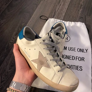 Golden Goose Superstar sneakers vit/blå - Golden Goose Superstar sneakers i vitt skinn med grå mockadetaljer och stjärna på sidan. Blå hälkappa i skinn, grå snörning och beige sula. Klassisk låg modell med rund tå och cool vintage vibe. Perfekta för dig som gillar streetstyle.