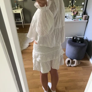 Vit volangkjol med prickar - Supersöt vit kortkjol med volanger och diskret prickigt mönster. Kjolen har bred smock i midjan för en bekväm passform och är gjord i ett lätt, luftigt tyg som passar perfekt till varma dagar.