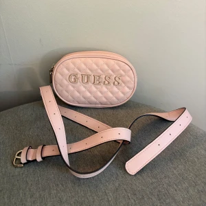 Ljusrosa quiltad axelväska från Guess - Supersöt ljusrosa axelväska från Guess med quiltad design, har justerbar rem och dragkedja. Insidan är fodrad med mönstrat tyg och har små fack för kort. Aldrig använd