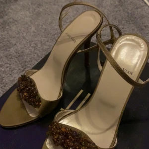 Guldiga sandaler från Venise Verte - Snygga sandaler i guldigt skinn från Venise Verte med smala remmar och hög klack. Framtill är de dekorerade med bruna pärlor som ger extra bling. Perfekta för dig som vill sticka ut med eleganta detaljer och öppen tå.