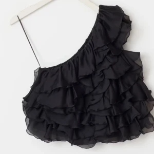 Svart organza one shoulder topp - Supersnygg svart topp med volanger och one shoulder-design. Tunt och lätt material som ger en luftig känsla. Perfekt för dig som vill sticka ut med en trendig och cool look. Volangerna ger extra attityd och toppen har en smal axelrem.
