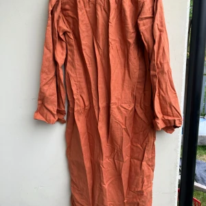 Lång orange linneklänning med brodyr - Säljer en långärmad klänning i en varm orange ton, sydd i linne. Klänningen har broderad detalj vid midjan och en rak, avslappnad passform. Längden är hellång och tyget är svalt och perfekt för varmare dagar. Klänning / Särk. STOR storlek. Ca 133 cm från halsringningen och till nerkant. Ca 140cm vid mätt precis under armarna.  Rostorange färg som är lite svår att fånga på bild. Dekorband vid hals och axlar. Snöre för stängning och reglering av vidd av halsringning.  Mycket material i den