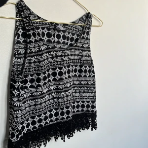 Svartvit blommig linnetopp med fransar - Säljer en svartvit linnetopp med blommigt mönster och snygga fransar längst ner. Toppen är ärmlös och har en lös passform, perfekt för varma dagar. Materialet känns lätt och luftigt, och mönstret ger en bohemisk vibe.