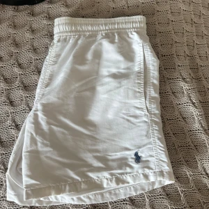 Vita badshorts från Polo Ralph Lauren - Snygga vita badshorts från Polo Ralph Lauren med elastisk midja och klassisk blå logga broderad framtill. Shortsen har sidfickor, bakficka och innerbyxa i mesh. Perfekta för stranden eller poolen under sommaren.