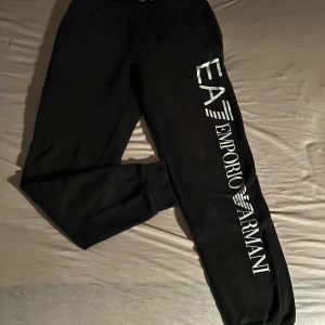 Svarta mjukisbyxor från Emporio Armani - Säljer ett par svarta mjukisbyxor från Emporio Armani med stort vitt tryck längs benet. Byxorna har en bekväm passform och resår i midjan samt vid bensluten. Perfekta för en avslappnad stil eller träning. 🖤