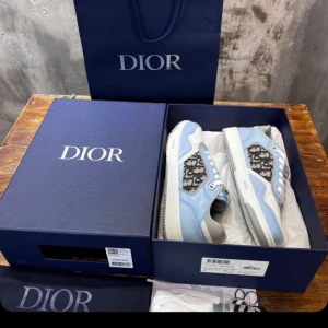 Dior sneakers i blått och vitt - Snygga Dior sneakers i ljusblått och vitt med klassiskt Dior-mönster på sidorna. Skorna har rund tå, snörning och platt sula. Perfekta för dig som vill sticka ut med exklusiv design och coola detaljer.