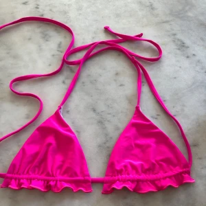 Rosa bikiniöverdel med volang - Supersöt bikiniöverdel i starkt rosa med volangkant och smala knytband. Klassisk trekantsmodell som ger en trendig och ungdomlig look, perfekt för stranden eller poolen. Fick den, det står ingen storlek men skulle tro det är en S.