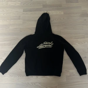 Svart Saint Laurent hoodie med tryck - Svart hoodie från Saint Laurent med ikoniskt logotyptryck i vitt framtill. Klassisk modell med huva och känguruficka. Tillverkad i mjuk bomull, perfekt för en avslappnad och trendig look. Långärmad och med ribbade muddar för extra komfort.