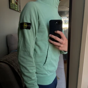 Mintgrön hoodie från Stone Island - Säljer en mintgrön hoodie från Stone Island med klassisk patch på vänster ärm. Tröjan har huva, känguruficka och är tillverkad i mjuk bomull. Perfekt för dig som vill uppgradera din garderob och vill ha en clean look med snygga detaljer. Den är storlek 156 men jag är 160 och den sitter bra.