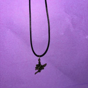 Svart halsband med fe-berlock - Ett svart halsband med en detaljrik fe-berlock i antikguld. Kedjan är tunn och smidig, perfekt för att ge en magisk touch till din stil. Berlocken har vackra vingar och en klassisk sagoinspirerad look.