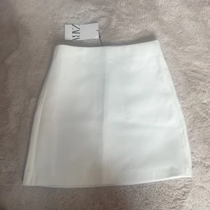 Vit satinliknande minikjol från Zara - Supersnygg vit kortkjol från Zara i satinliknande material. Kjolen har en stilren och enkel design med slät yta och lätt glans. Perfekt för dig som gillar minimalistisk och fräsch stil. Kjolen har en rak passform och är fodrad för extra komfort.