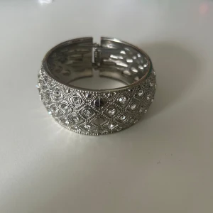 Bredt silverfärgat armband med stenar - Stilrent och chunky armband i silverfärg med glittrande stenar infattade i ett rutmönster. Armbandet har en bred design och stängs med ett smidigt gångjärn. Perfekt statement piece för dig som gillar att sticka ut.