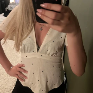 Prickig v-ringad blus i ljus beige - Supersöt ljusbeige blus med svarta prickar och djup v-ringning. Toppen har korta ärmar och band på sidorna som man kan knyta baktill. 