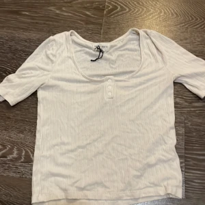 Vit ribbad topp Holly & Whyte S - Vit ribbad topp från Holly & Whyte by Lindex i storlek S passar även M. Modellen har korta ärmar, rundad djup ringning och tre dekorativa knappar framtill. Perfekt för en clean och enkel look.