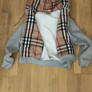 Grå hoodiejacka från Burberry - Grå hoodiejacka från Burberry med klassiskt rutigt foder i beige, svart, rött och vitt. Jackan har dragkedja, snörning i huvan och mjukt vitt innerfoder. Perfekt för dig som vill ha en stilren men ändå lyxig look.