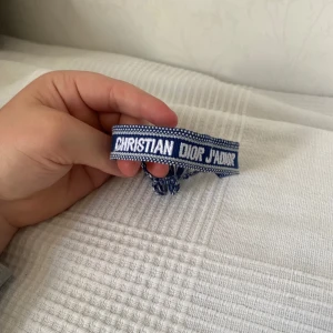 Blått armband Christian Dior J'Adior - Snyggt blått armband från Christian Dior med texten 'J'Adior' broderad i vitt. Armbandet har justerbar knytning och coola tofsar i blått och vitt. Perfekt accessoar för att lyfta din outfit med en lyxig touch.