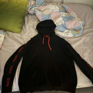 Svart hoodie från Hugo med röda detaljer - Svart hoodie från Hugo med röda snören och röd logga längs båda ärmarna. Klassisk huva och känguruficka framtill. Snygg streetstil som sticker ut med de tydliga loggdetaljerna. Perfekt för dig som gillar en clean men ändå bold look. Nypris - 2000kr