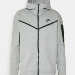 Grå zip-hoodie från Nike - Snygg grå zip-hoodie från Nike med svart logga på bröstet och svarta detaljer längs dragkedjan och över bröstet. Jackan har huva, lång ärm och en extra ficka med dragkedja på ena ärmen. Perfekt för en sportig och avslappnad stil.