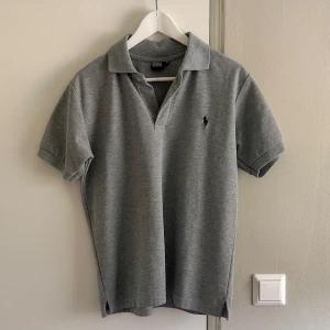 Grå pikétröja från Polo Ralph Lauren - Klassisk grå pikétröja från Polo Ralph Lauren i 100% bomull. Tröjan har korta ärmar, krage och den klassiska loggan i mörkblått på bröstet. Perfekt basic med snygg passform och tidlös stil. Passar perfekt nu till sommaren.