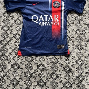 PSG Mbappé matchtröja Nike blå - Paris Saint-Germain fotbollströja med Mbappé 7 på ryggen. Djupblå med röda och vita detaljer, PSG-emblem på bröstet och Qatar Airways-tryck. Tillverkad i lätt, ventilerande material med korta ärmar och rund hals. Snyggt mönster i tyget och Nike-logga i rött.