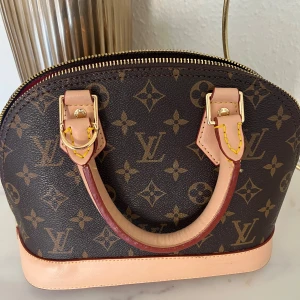 Alma bb handväska från Louis Vuitton - Elegant handväska i klassiskt monogrammönster från Louis Vuitton. Väskan har en dragkedja upptill och detaljer i ljusbrunt skinn. Perfekt för att ge en lyxig touch till din outfit.