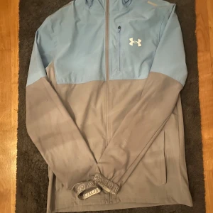 Under Armour vindjacka blå/grå - Snygg vindjacka från Under Armour i ljusblått och grått. Jackan har huva, hel dragkedja, bröstficka med dragkedja och logga på bröstet. Perfekt för dig som gillar sportig stil och vill ha något lätt och funktionellt.