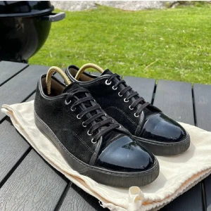 Svarta sneakers Lanvin - Stilrena svarta sneakers med ovandel i mocka och snygg lackad tå. Skorna har snörning och en platt sula, vilket ger en clean och modern look. Perfekta för dig som gillar en klassisk men ändå edgy stil.