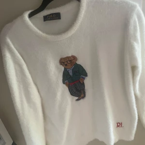 Vit fluffig tröja Polo Ralph Lauren - Supermjuk vit tröja från Polo Ralph Lauren med ikonisk nallebrodyr på bröstet. Tröjan har lång ärm, rund hals och RL-broderi nertill. Perfekt för dig som gillar en mysig och stilren look med en twist. Passar xs/s. 